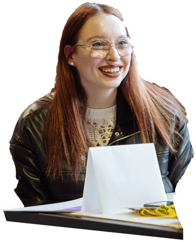 Une jeune femme aux longs cheveux roux et aux grandes lunettes sourit à son interlocuteur, derrière une petite table.